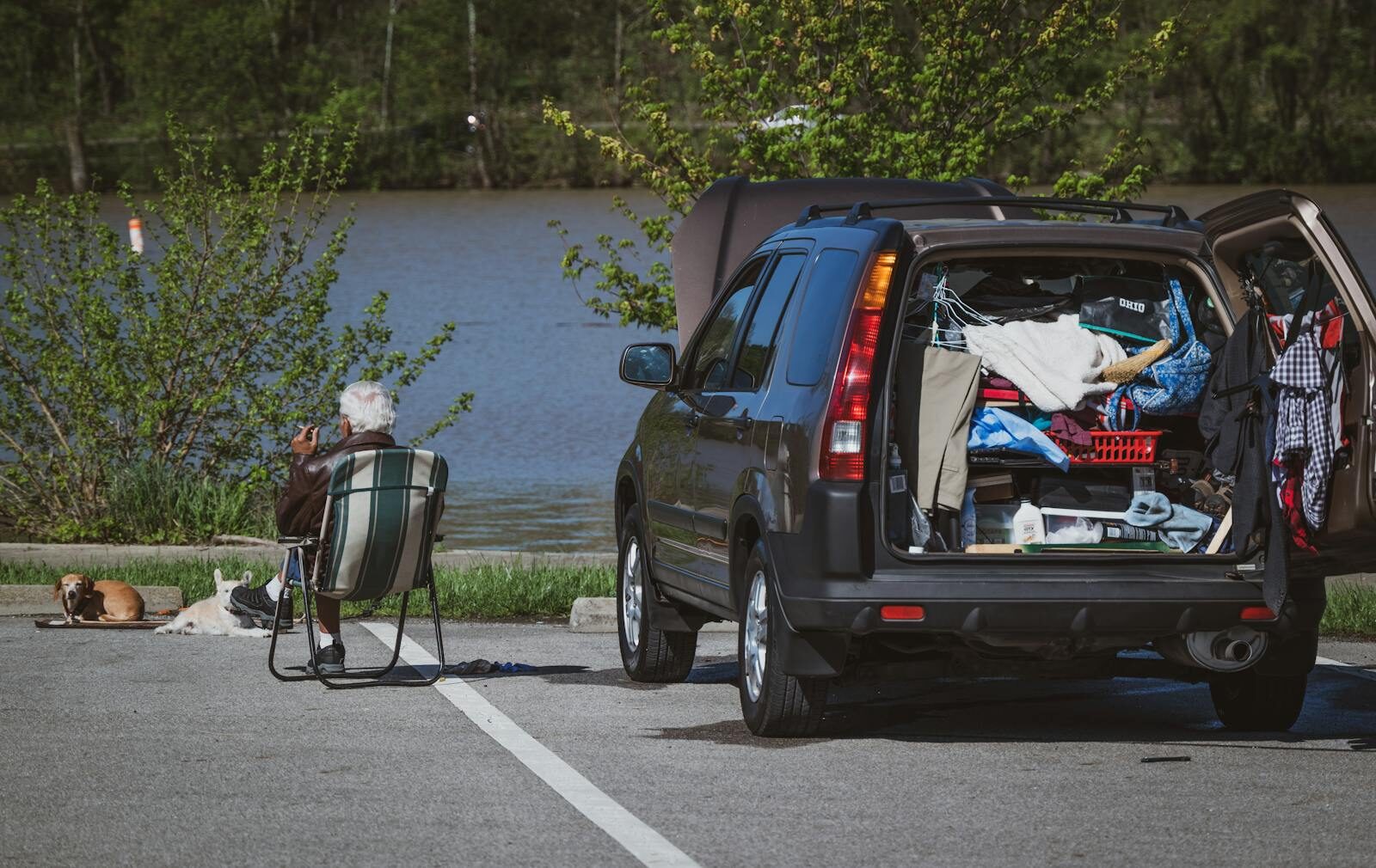 An elderly man sits beside his open car trunk filled with belongings, enjoying a riverside view.