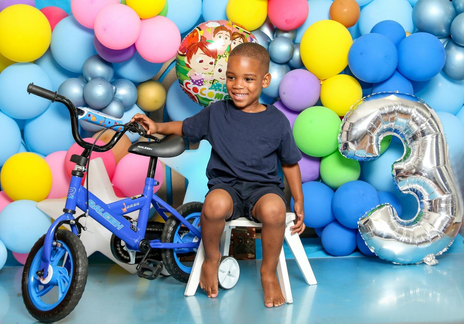 Smiling boy celebrates third birthday with balloons and a new blue bicycle.
