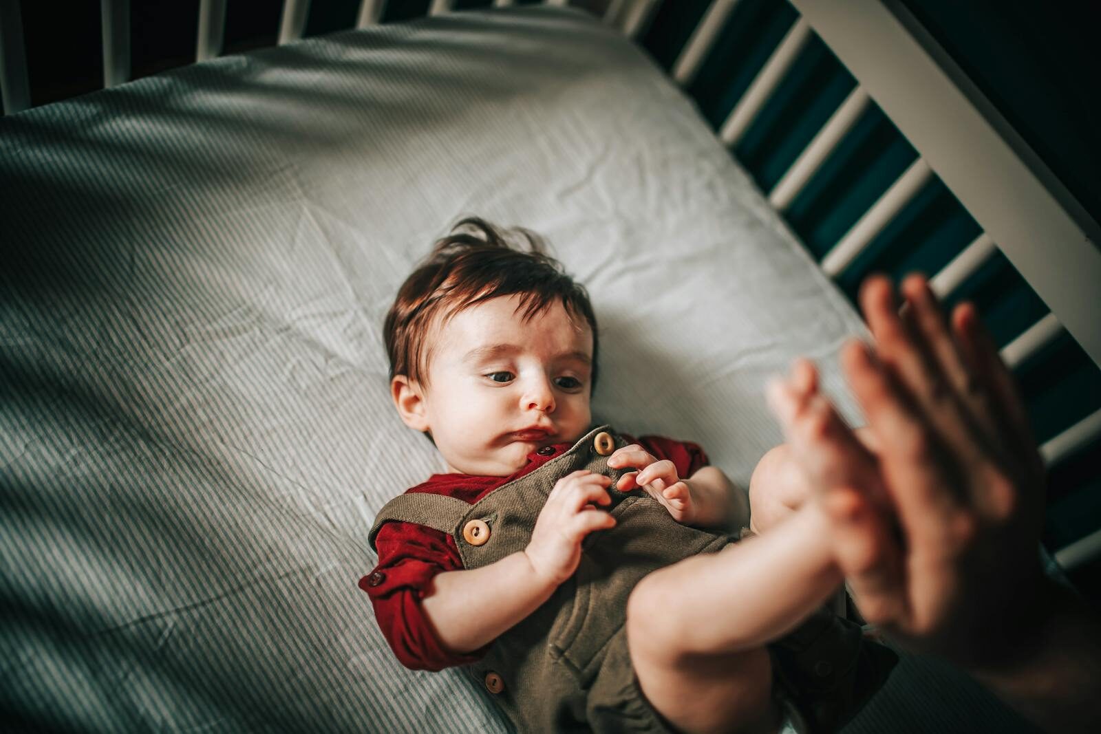 From above of crop anonymous parent touching feet of adorable little boy lying in crib on crumpled sheet while looking away at home