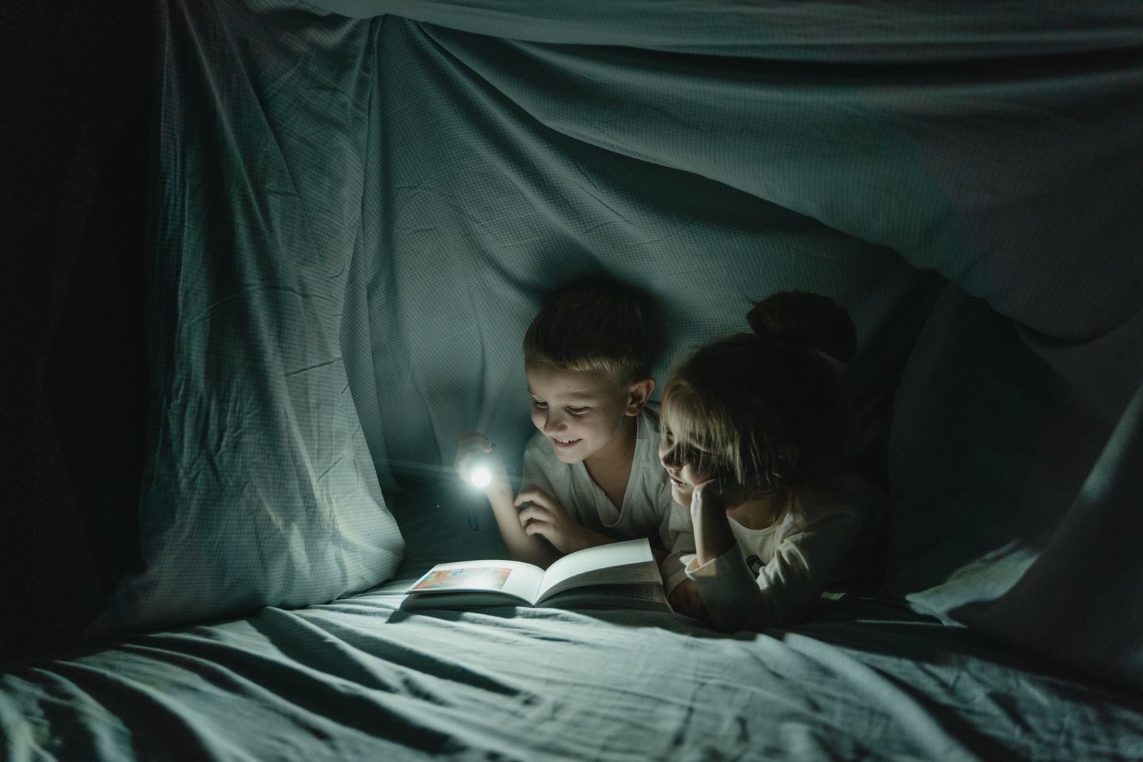 Two children enjoy reading under a cozy blanket fort, illuminated by a flashlight, creating a warm nighttime atmosphere.