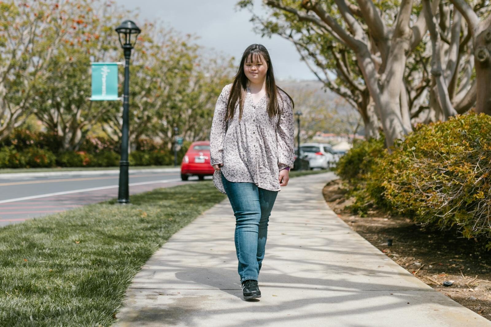 Young woman with long hair walks on a sidewalk lined with trees, wearing a floral top and jeans.