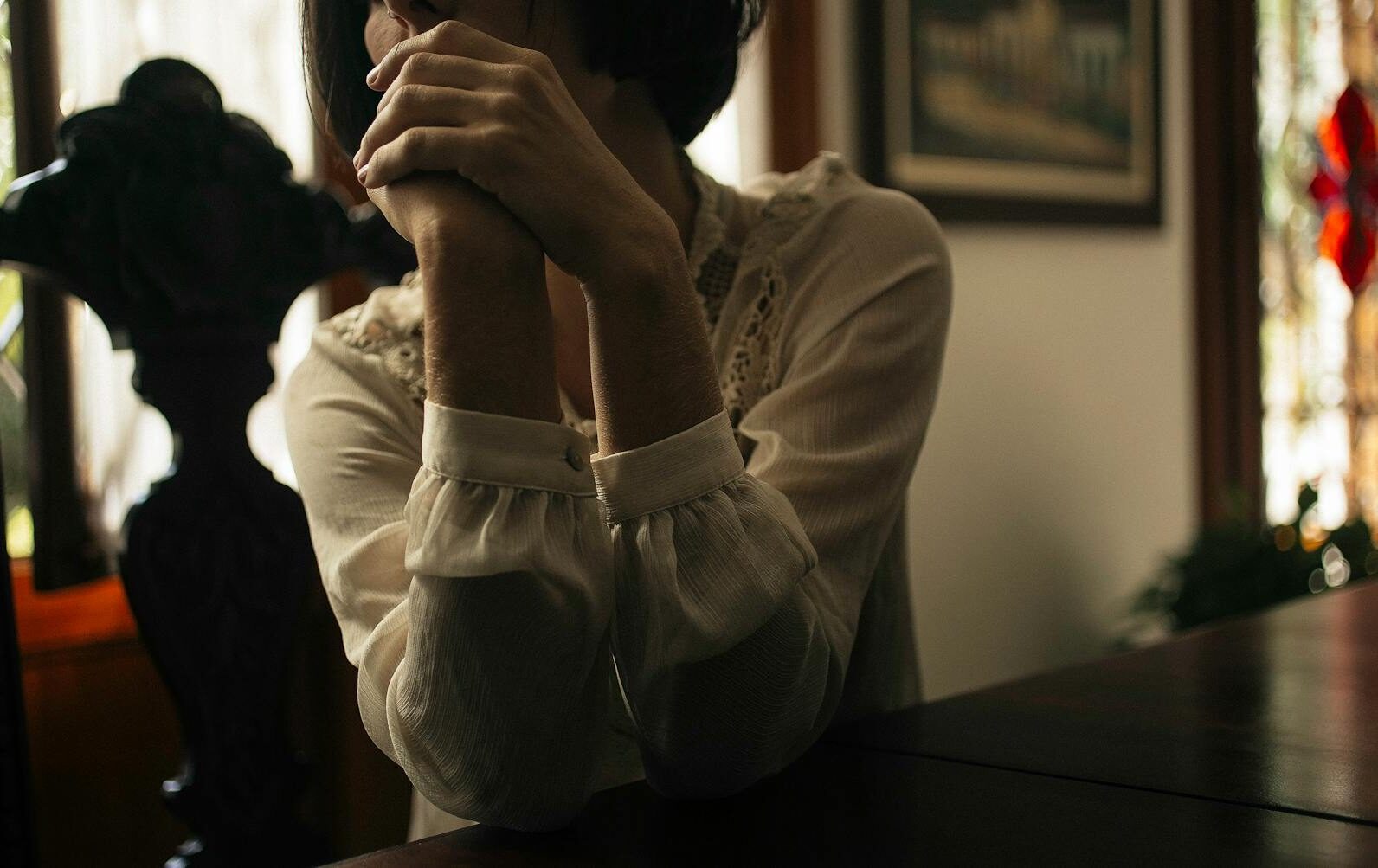 Pensive woman sitting at a table, hands clasped, indoors with soft lighting.