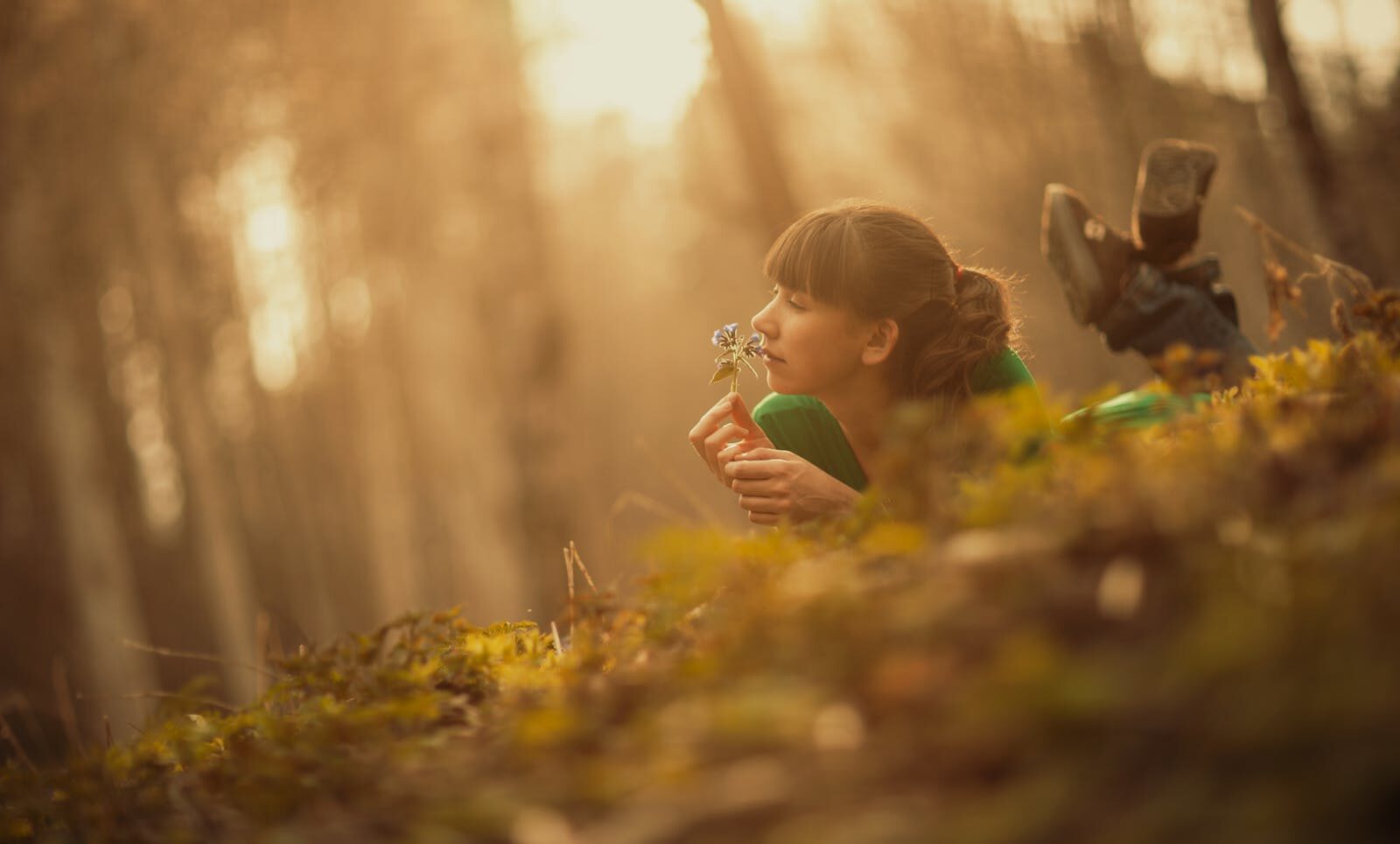 A woman lies in the forest, enjoying the scent of a wildflower, surrounded by warm sunlight.